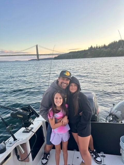 Captain Ryan Montoya with his two daughters on a fishing boat near Puget Sound bridge at sunset.